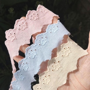 May include: Three strips of scalloped eyelet lace trim in pastel pink, light blue, and cream. Each strip features a repeating embroidered pattern of small paw prints along the scalloped edge, with delicate circular cutouts and stitching.