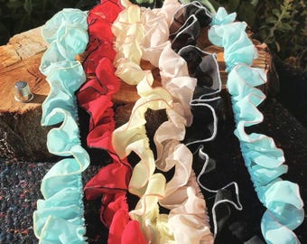 May include: A collection of five ruffled fabric trims in light blue, red, pale yellow, beige, and black. Each strip features a gathered, wavy edge with contrasting thread stitching along the border. A small metal thimble sits to the left.