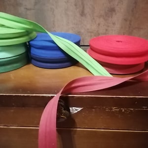 Könnte beinhalten: Vier Spulen mit Schrägband in verschiedenen Farben: grün, blau, rot und burgunderrot. Das Band ist in der Mitte gefaltet und bereit für Nähprojekte.