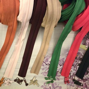 Könnte beinhalten: Eine Sammlung farbenfroher Reißverschlüsse in verschiedenen Farbtönen, darunter Orange, Rosa, Burgunder, Beige, Grün, Rot und Schwarz. Die Reißverschlüsse werden mit den passenden Zippern präsentiert und zeigen eine Auswahl an Nähzubehör.