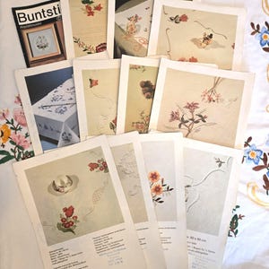 Könnte beinhalten: Eine Sammlung von Vintage-Stickmustern mit floralen Designs und Tischdeckenmotiven. Die Blätter sind auf einem weißen, floral bestickten Stoff angeordnet. Ein Heft mit dem Titel "Buntsti" ist ebenfalls sichtbar.