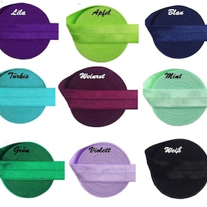 Könnte beinhalten: Neun Rollen elastisches Stoffband in verschiedenen Farben, darunter Lila, Grün, Blau, Türkis, Weinrot, Mint, Grün, Violett und Schwarz. Jede Rolle hat ein Etikett mit einem deutschen Wort für die Farbe.