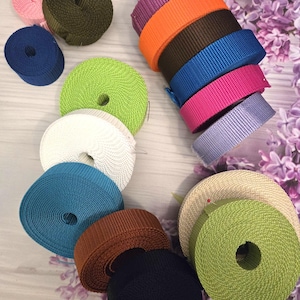 Può includere: Una collezione di rotoli colorati di fettuccia di nylon in varie tonalità, tra cui rosa, verde, blu e arancione. I rotoli sono disposti ordinatamente, mostrando i diversi colori e le trame del materiale. La fettuccia è probabilmente utilizzata per l'artigianato o l'attrezzatura da esterno.