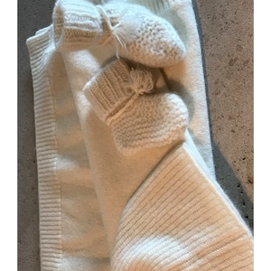 May include: Two pairs of white knitted baby mittens with pom-poms. The mittens are folded on top of a white knitted sweater.