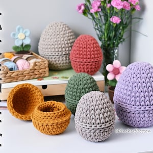 Może przedstawiać: Kolekcja dzianych koszyków w kształcie jajek w różnych kolorach, w tym fioletowym, szarym, zielonym, czerwonym i beżowym. Koszyki są ułożone na białej powierzchni z drewnianą półką w tle. Tekst "aPinchOfCotton" jest widoczny w prawym dolnym rogu obrazu.