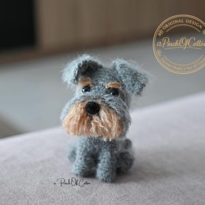 Può includere: Una piccola statuetta di cane all'uncinetto di colore grigio con un aspetto testurizzato e soffice. Il cane ha un muso color marrone chiaro, occhi a bottone neri e un naso nero. Un logo circolare in alto a destra riporta Original Design aPinchOfCotton Human Made I No AI Used.