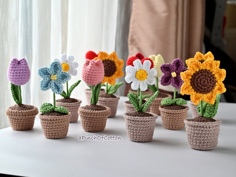 May include: A collection of crocheted flower pots in various colors, including purple, blue, pink, white, yellow, and brown. The flowers are arranged in a row on a white surface. The text "aPinchOfCotton" is visible in the bottom right corner of the image.