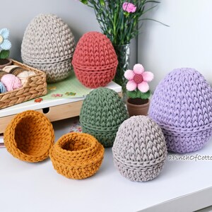 May include: A collection of crocheted egg-shaped baskets in various colors, including purple, gray, green, and orange. The baskets are arranged on a white surface with a wicker basket filled with yarn in the background.