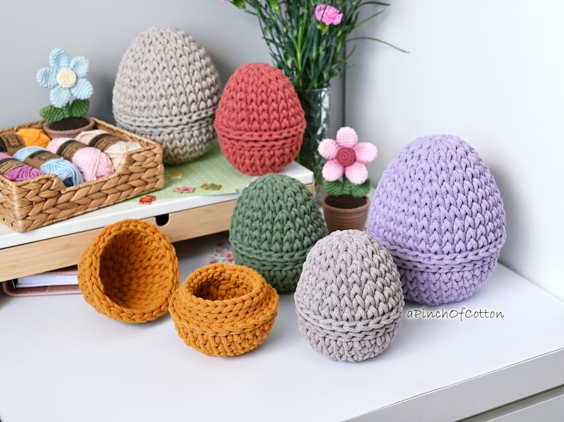 May include: A collection of crocheted egg-shaped baskets in various colors, including purple, gray, green, and orange. The baskets are arranged on a white surface with a wicker basket filled with yarn in the background.
