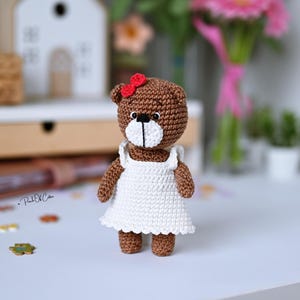 May include: A handmade brown crochet bear wearing a white dress and a red bow. The bear has a white muzzle and black eyes. The bear is standing on a white surface. The bear is a handmade item.