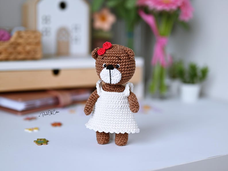 May include: A handmade brown crochet bear wearing a white dress and a red bow. The bear has a white muzzle and black eyes. The bear is standing on a white surface. The bear is a handmade item.