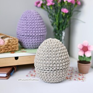 May include: Two crocheted egg-shaped baskets, one in light purple and one in beige, on a white surface with a wicker basket filled with yarn in the background.