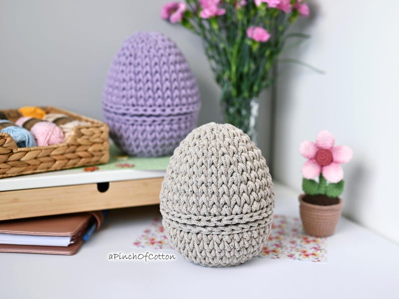 May include: Two crocheted egg-shaped baskets, one in light purple and one in beige, on a white surface with a wicker basket filled with yarn in the background.