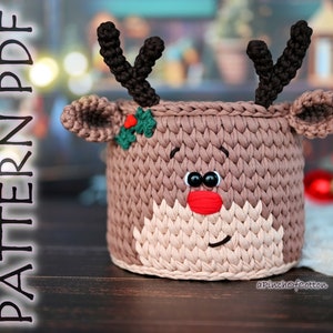May include: A crocheted reindeer-shaped basket in brown and beige tones. The basket has a red nose, black eyes, and a green holly sprig on its head. The text "PATTERN PDF" is visible on the left side of the image.