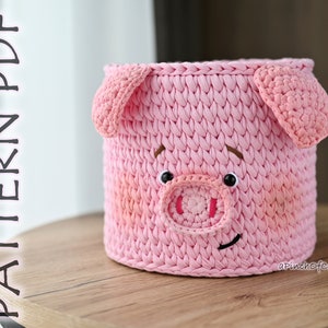 May include: Pink crocheted pig-shaped basket with a smiling face. The basket is made with a textured stitch and has a white background with pink accents. The basket is sitting on a wooden surface.