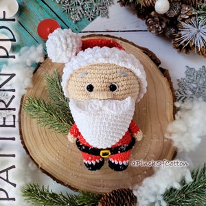 May include: Crocheted Santa Claus amigurumi doll with a white beard, red hat, and red and white suit. The doll is sitting on a wooden slice with a white background.