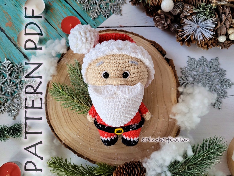 May include: Crocheted Santa Claus amigurumi doll with a white beard, red hat, and red and white suit. The doll is sitting on a wooden slice with a white background.