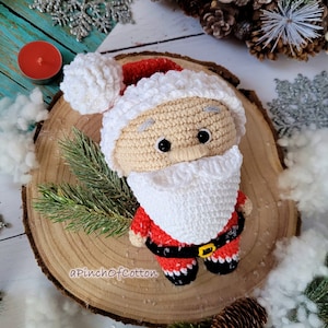May include: Crocheted Santa Claus doll with a white beard, red suit, and black belt. The doll is sitting on a wooden slice with a white background.