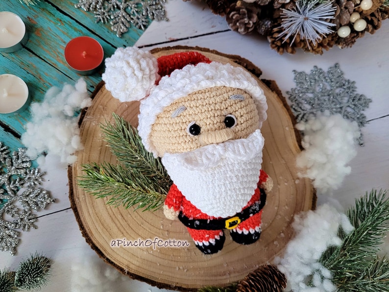 May include: Crocheted Santa Claus doll with a white beard, red suit, and black belt. The doll is sitting on a wooden slice with a white background.