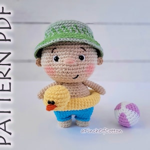 Può includere: Bambola amigurumi all'uncinetto di un bambino che indossa un cappello a secchiello verde, pantaloncini da bagno blu e un salvagente gonfiabile giallo. La bambola tiene un anatroccolo di gomma giallo e una palla da spiaggia rosa e bianca all'uncinetto.