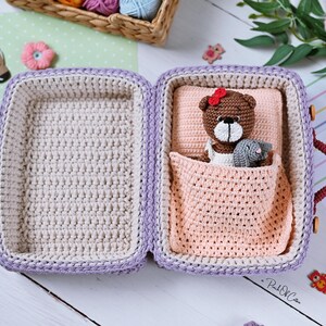 May include: Crocheted suitcase in lavender and cream, open to reveal a bed with a brown teddy bear wearing a red bow and a gray bunny. The suitcase has burgundy handles and wooden closures. Decorative flowers and a basket of yarn are in the background.