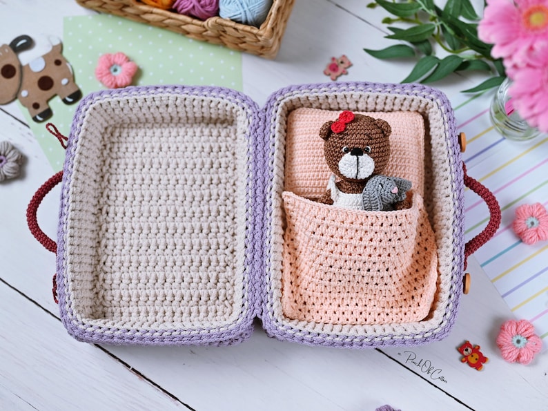 May include: Crocheted suitcase in lavender and cream, open to reveal a bed with a brown teddy bear wearing a red bow and a gray bunny. The suitcase has burgundy handles and wooden closures. Decorative flowers and a basket of yarn are in the background.