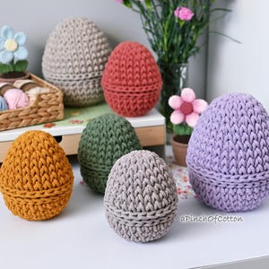 May include: Six crocheted egg-shaped containers in various colors, including purple, gray, green, orange, and red. The containers are arranged on a white surface with a wooden shelf in the background.