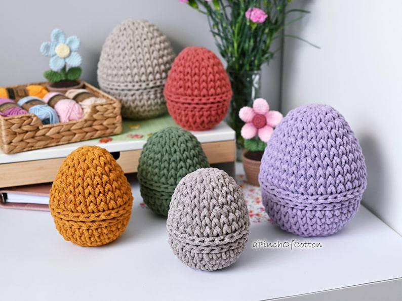 May include: Six crocheted egg-shaped containers in various colors, including purple, gray, green, orange, and red. The containers are arranged on a white surface with a wooden shelf in the background.