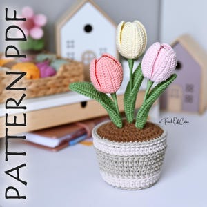 May include: A crocheted tulip arrangement in a striped pot. The arrangement features three tulips in pink, yellow, and pink, with green leaves. The pot is a mix of brown, gray, and white. The text "PATTERN PDF" is on the left side of the image.