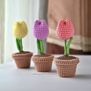 May include: Three crocheted tulip flowers in pots. The tulips are yellow, pink, and purple. The pots are brown.