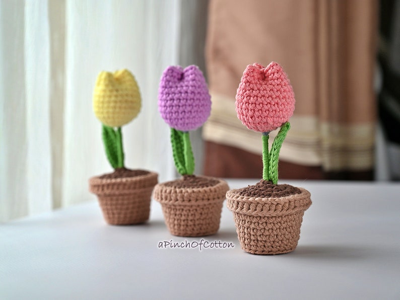 May include: Three crocheted tulip flowers in pots. The tulips are yellow, pink, and purple. The pots are brown.