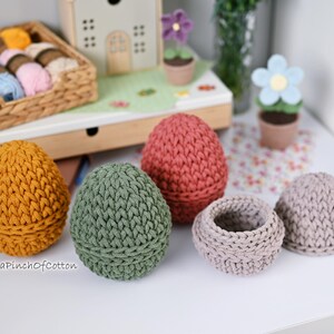 May include: Five crocheted egg-shaped containers in different colors, including green, brown, pink, and gray. The containers are made with a textured stitch and are perfect for storing small items.