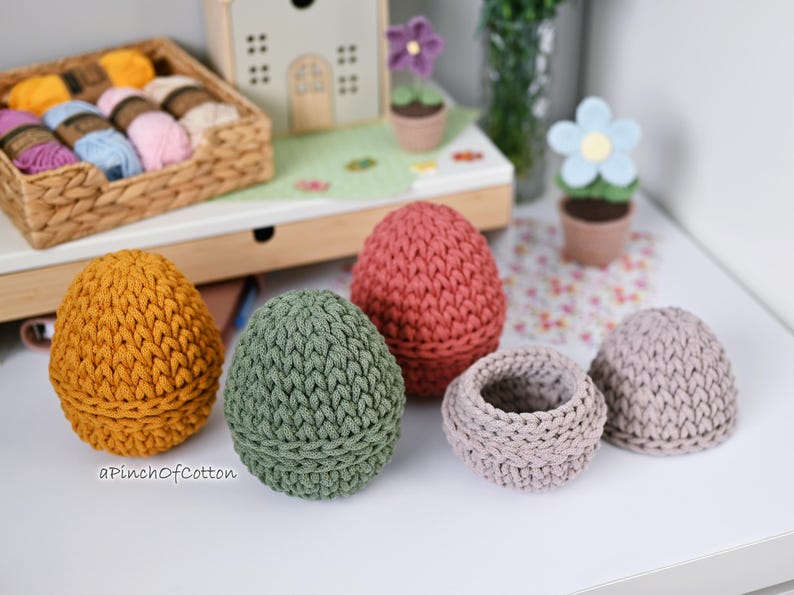 May include: Five crocheted egg-shaped containers in different colors, including green, brown, pink, and gray. The containers are made with a textured stitch and are perfect for storing small items.