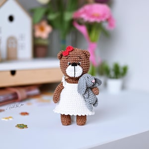 May include: A handmade brown crochet bear wearing a white dress and a red bow, holding a gray crochet bunny. The bear and bunny are on a white surface, with a blurred background featuring flowers and a small wooden house.