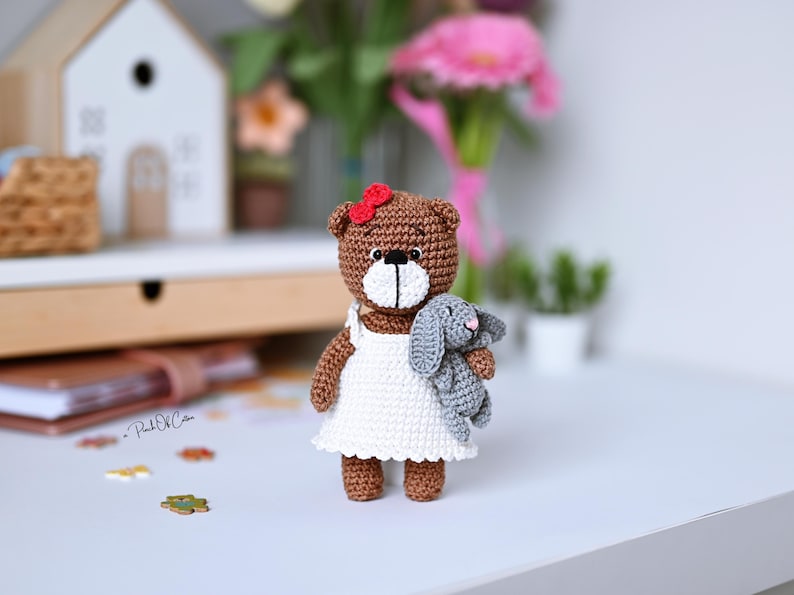 May include: A handmade brown crochet bear wearing a white dress and a red bow, holding a gray crochet bunny. The bear and bunny are on a white surface, with a blurred background featuring flowers and a small wooden house.