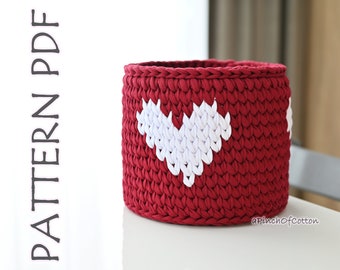 May include: A red crocheted basket with a white heart design. The basket is sitting on a white surface.