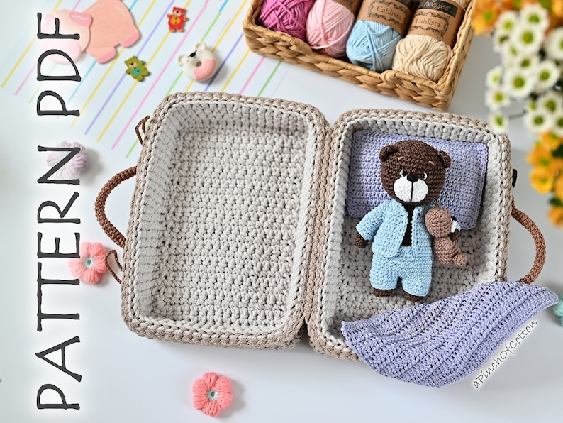 K&ouml;nnte beinhalten: Ein geh&auml;kelter Koffer mit einem braunen Griff, einem braunen Teddyb&auml;r in blauem Schlafanzug, einer lila Decke und einem kleineren braunen Teddyb&auml;r. Der Koffer ist ge&ouml;ffnet und der Teddyb&auml;r befindet sich im Inneren. Der Text "a Pinch Of Cotton" ist in der rechten unteren Ecke des Bildes sichtbar.