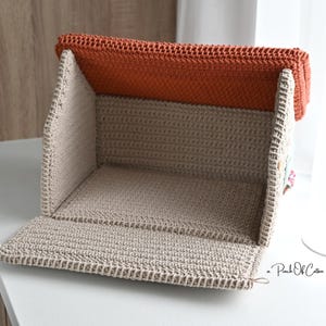 以下が含まれることがあります： ベージュとオレンジ色の手編みの収納ボックス。ボックスは開いており、内部が見えます。上部はオレンジ色で、残りのボックスはベージュ色です。ボックスは白い表面に置かれています。