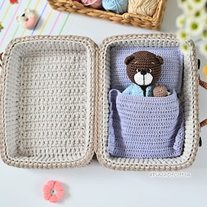 K&ouml;nnte beinhalten: Ein geh&auml;kelter Koffer mit einem braunen Teddyb&auml;ren im Inneren. Der Koffer ist ge&ouml;ffnet und der Teddyb&auml;r liegt in einer lila Decke. Der Text "aPinchOfCotton" ist in der unteren rechten Ecke des Bildes sichtbar.