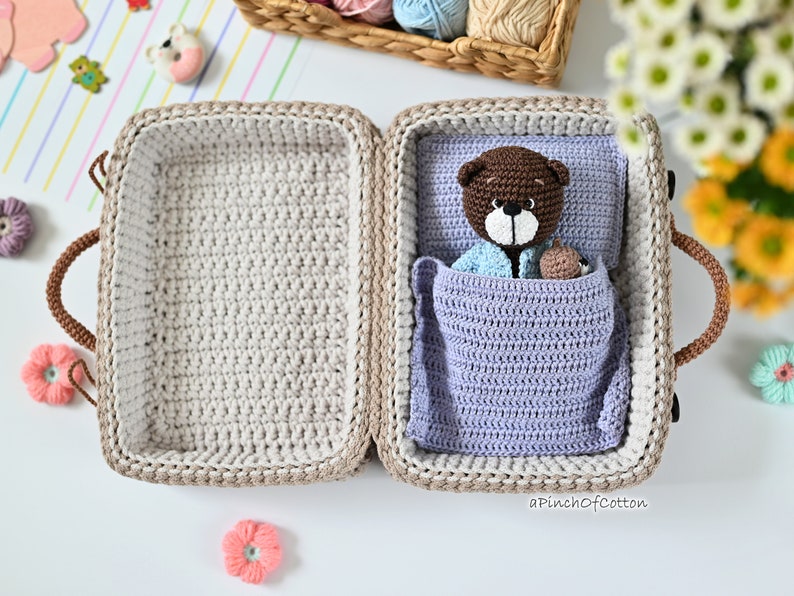 K&ouml;nnte beinhalten: Ein geh&auml;kelter Koffer mit einem braunen Teddyb&auml;ren im Inneren. Der Koffer ist ge&ouml;ffnet und der Teddyb&auml;r liegt in einer lila Decke. Der Text "aPinchOfCotton" ist in der unteren rechten Ecke des Bildes sichtbar.
