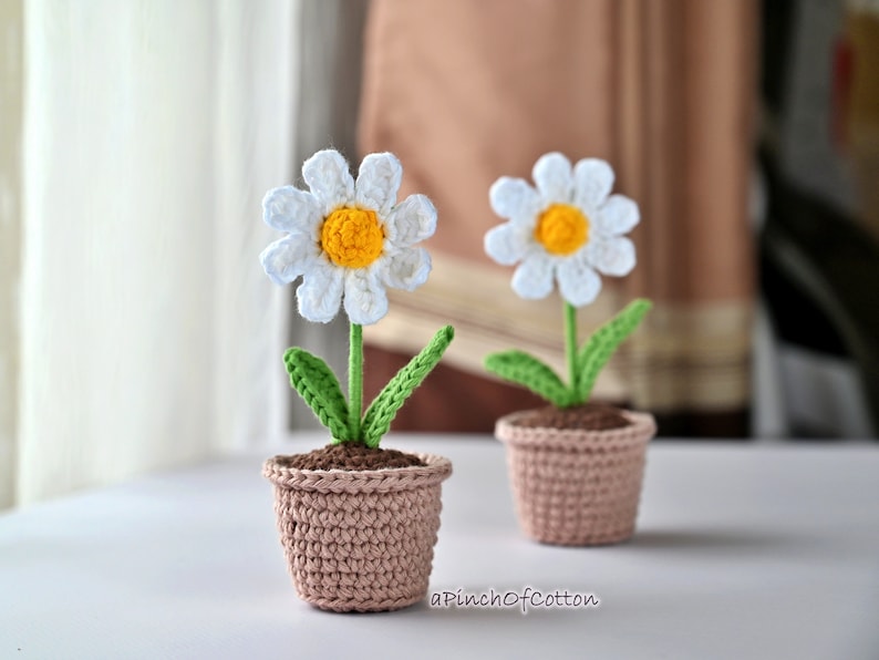 May include: Two crocheted daisy flowers in brown pots. The daisies have white petals, yellow centers, and green leaves. The pots are made of brown yarn.