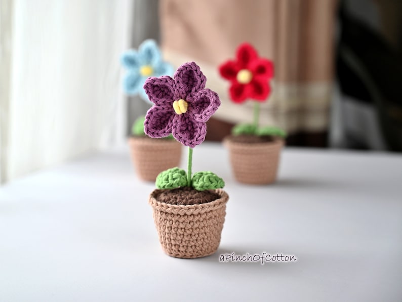 May include: Three crocheted flower pots with different colored flowers. The flowers are purple, blue, and red. The pots are brown and the flowers are sitting on a white surface.