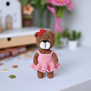 May include: A handmade crochet teddy bear with a brown body, white muzzle, and a red bow. The bear wears a pink dress. The bear is standing on a white surface. The background includes a white house, flowers, and other decorative items.