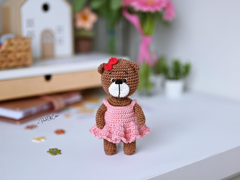 May include: A handmade crochet teddy bear with a brown body, white muzzle, and a red bow. The bear wears a pink dress. The bear is standing on a white surface. The background includes a white house, flowers, and other decorative items.