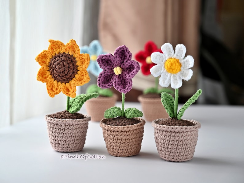 May include: Four crocheted flower pots with different colored flowers. The flowers are a sunflower, a purple flower, a white flower, and a red flower. The pots are all brown and have a crocheted texture.