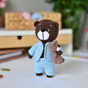 K&ouml;nnte beinhalten: Ein brauner H&auml;kelteddyb&auml;r, der ein hellblaues H&auml;keloutfit tr&auml;gt. Der B&auml;r h&auml;lt einen kleineren braunen H&auml;kelteddyb&auml;ren. Die B&auml;ren befinden sich auf einer wei&szlig;en Oberfl&auml;che.