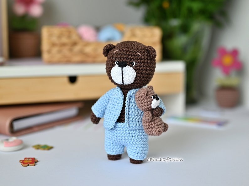 K&ouml;nnte beinhalten: Ein brauner H&auml;kelteddyb&auml;r, der ein hellblaues H&auml;keloutfit tr&auml;gt. Der B&auml;r h&auml;lt einen kleineren braunen H&auml;kelteddyb&auml;ren. Die B&auml;ren befinden sich auf einer wei&szlig;en Oberfl&auml;che.