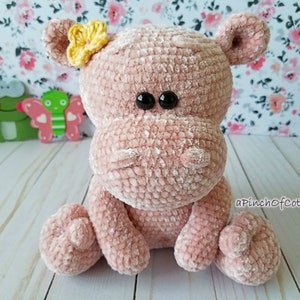 May include: A plush, pink crocheted hippo with black button eyes and a yellow flower on its head. The handmade hippo is sitting with its legs extended. The text "aPinchofCotton" is visible on the lower right.