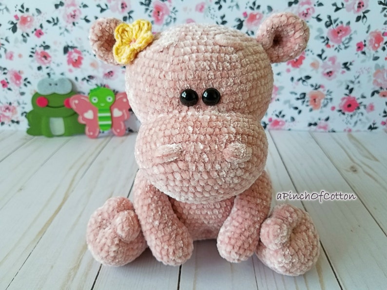 May include: A plush, pink crocheted hippo with black button eyes and a yellow flower on its head. The handmade hippo is sitting with its legs extended. The text "aPinchofCotton" is visible on the lower right.