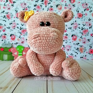 May include: A handmade, plush pink hippo with black eyes and a yellow bow. The hippo is crocheted and sitting upright. The background features a floral pattern and a wooden surface. The text "apinchofcotton" is visible.